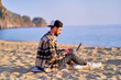 © Goffkein - Young happy joyful carefree satisfied millennial freelancer man using laptop on sand beach by the sea. Dream office work concept