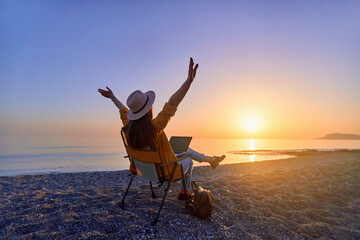 Satisfied free millennial freelancer woman with open arms using computer and sitting on the beach by the calm sea. Enjoyment of dream office remote work concept