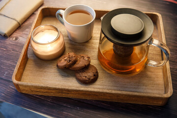  Interior and home cosiness concept. Top view. Burning candle in a jar, a cup of tea, a teapot with herbal tea and three oatmeal chocolate chip cookies on a wooden tray on the table