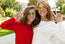 Stylish Happy Two Girls In Bright Round Sunglasses Smiling And Showing Peace Sigh At Camera. Positive Lovely Girls Smiling On City Background.