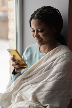 Smiling black woman with yellow cellphone sitting on windowsill at home and makes purchase in online store. African American young female with warm blanket texting with her friends on ledge by window 