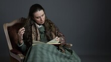 A Victorian Woman With Long, Dark Hair And Wearing A Green Checked Dress And A Paisley Shawl Sitting In A Chair Reading A Book
