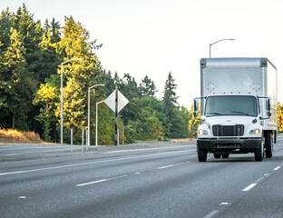 Day cab white middle powered semi truck with box trailer running on the highway road with local entrance and autumn trees on the side