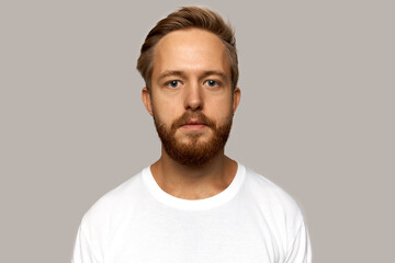 Wall Mural - Horizontal picture of attractive Caucasian cute bearded guy posing in white cotton t-shirt against grey background of studio wall looking at camera, having stylish haircut and calm face expression