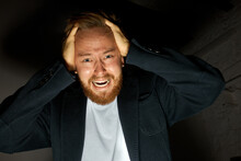 Low-key Light Photo Of Desperate Furious And Angry Man In Dark At Studio, Holding Head And Screaming With Red Face, Dressed In Black Coat And White T-shirt, Having Scared Face Expression