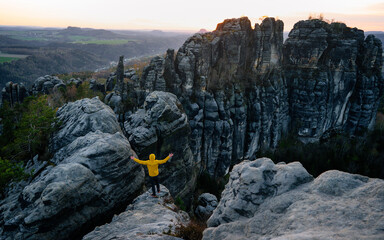 Young man standing on a cliff edge celebrates reaching the top of the mountain, arms in the air, shouting.