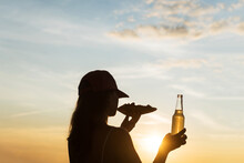 Silhouette Of Girl In Baseball Cap Eating Pizza And Drinking Soda Water From Glass Bottle And Looking At Sunset Sky. Rear View Of Female
