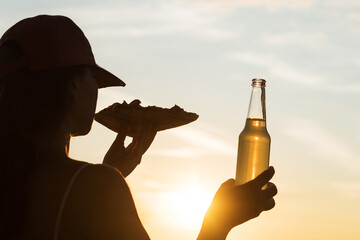 Silhouette of girl in baseball cap eating pizza and drinking soda water from glass bottle and looking at sunset sky. Rear view of female