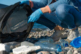 Fototapeta Uliczki - Young woman is squatting and collecting plastic, rubber garbage, foam in a black garbage bag on a pebble beach by the sea. Concept of ecology, volunteerism, kindness