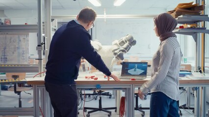 Leinwandbilder - Senior Male Engineer Explaining to Muslim Female Computer Science Specialist and Showing at Robotic Hand at the Research Laboratory with Laptop. Innovative Firm Startup Concept