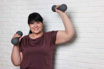 Canvas Print - Happy overweight mature woman doing exercise with dumbbells near white brick wall