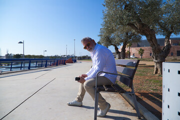 Handsome mature man with beard and grey hair is sitting thoughtfully on a park bench while consulting his mobile phone. The man is dressed in beige trousers, white shirt. Concept of old age.