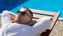 Portrait Of A Relaxed Young Man In Sunglasses And A White Coat, Sunbathing By The Pool With His Hands Behind His Head. Close-up.