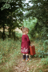 Rear view of a pretty little girl in burgundy boho dress holding her old suitcases in the forest at summer time, retro style