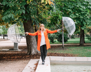 blonde girl with blue eyes and dressed in red gabardine balances with two transparent umbrellas in a park in Madrid, Spain.