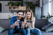 © Liubomir - Happy couple man and woman playing game using joystick while sitting on sofa at home