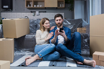 A man and a woman in a new apartment, a married couple sitting on the floor, near cardboard boxes, using the phone for online shopping, choosing furniture