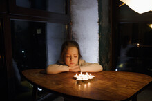 A Little Girl With Her Eyes Closed Is Sitting At A Table In Front Of Burning Candles. The Child Makes A Wish