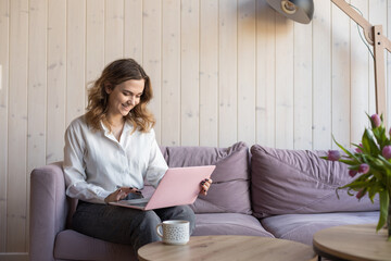 Portrait of smiling woman working on pink laptop sitting on soft violet sofa in casual clothes near table with cup of coffee. Living room in elegant design with aesthetic decor, free copy space