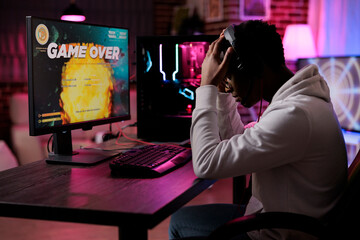 Wall Mural - Frustrated young adult losing online video games play competition on computer with neon lights. Male gamer streaming action gameplay and feeling sad about lost shooting championship.