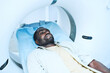 Serious young black patient with stubble lying on table while moving into MRI scanner in clinic