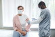 © Mediaphotos - Concentrated African-American nurse in mask giving covid vaccine to patient on couch in hospital