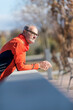 © 2Design - Front view of a senior runner man with sportswear sitting on a wooden fence while looking away