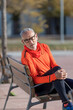 © 2Design - Front view of senior man in sports clothes sitting in a bench at park while smiling and looking camera in a sunny day
