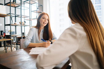 Wall Mural - Back view of two smiling business woman working together on contract documents sitting at desk with laptop in modern office room. Happy female coworkers meeting for new project planning in boardroom.