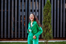 Cheerful Smiling Brunette Woman In Green Suit Posing Next To Black Rack Wall, Urban Building.