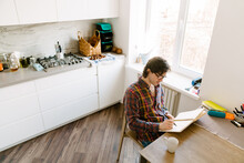 White Man Wearing Glasses Writing Down Notes While Sitting In Kitchen