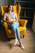© dikushin - Vertical portrait of attarctive blonde woman resting in comfortable yellow chair holding paper book in dark room with modern interior, smiling looking at camera, on background of bookshelves.