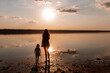 Young mother walks on the water with daughter in straw hat at sunset. Girls walk barefoot on the sea
