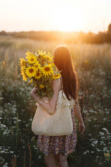Wall Mural - Beautiful woman gathering sunflowers in warm sunset light in summer meadow. Tranquil atmospheric moment in countryside. Stylish young female in floral dress holding sunflowers in evening field