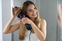 Portrait Of Young Woman Using Round Brush Hair Dryer To Style Hair At The Mirror In An Easy Way At Home