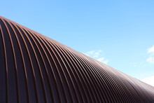 The Roof Of The Hangar Is Made Of Stainless Steel. Arched Roof Made Of Metal Construction. An Industrial Building In Detail Against A Blue Sky Background.