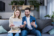 © Liubomir - A man and a woman are happy at home, make online payments, using the phone and banking application, sitting on the couch and holding bills