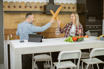 Funny middle aged couple using fresh baguettes for playful fight on bright kitchen. Caucasian family of two loving people spending time at home with fun and pleasure.