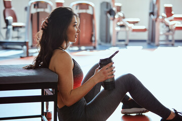 Wall Mural - A sportswoman sits on the floor in a gym and drinks a protein shake for strength.