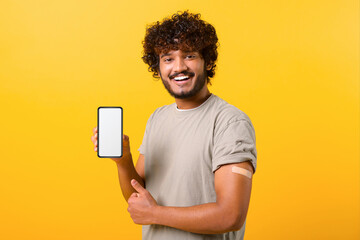 Smiling curly Indian man with medical band-aid on arm after the vaccination showing smartphone with empty screen isolated on yellow, demonstrate international certificate, qr code, copy space, mockup