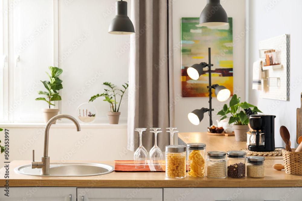 Wooden counter with silver sink, glasses and jars in light kitchen