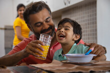 Close-up Portrait Of Caring Father Feeding Orange Juice To His Little Child And Mother Standing Behind