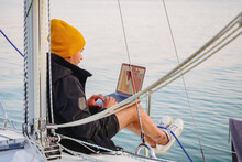 Handsome Young Person Wear Jacket And Knitted Hat Working With Laptop On A Sailing Yacht During Sailing By Lake, Remote Work Concept