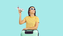 Happy Woman Traveler In Sunglasses With Suitcase Hold Airplane Model In Hands Isolated On Green Studio Background. Smiling Girl With Baggage Ready For Summer Vacation. Travel And Tourism.