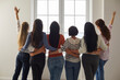 © Studio Romantic - Back rear view of group of happy confident beautiful slim young women in their 20s, 30s and 40s standing together, in row, hugging each other around waist and looking forward through big bright window
