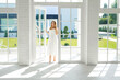 © Maryna - A young beautiful elegant woman enters the open door of a house with huge high panoramic windows to the floor, through which rays of sunlight from the street will come. Clean bright white room