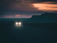 Van Driving On A Road In The Utah Desert Landscape At Sunset. Moody Clouds Darken The Skies. Vanlife At Twilight. Headlights Shining With A Butte In The Background. Traveling The American Southwest