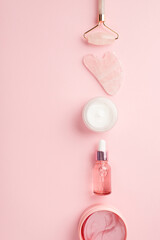 Wall Mural - Beauty procedure concept. Top view vertical photo of rose quartz roller gua sha pink eye patches glass dropper bottle with serum and cream jar on isolated pastel pink background