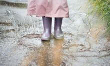 Little Girl In Pink Waterproof Raincoat, Purple Rubber Boots Funny Jumps Through Puddles On Street Road In Rainy Day Weather. Spring, Autumn. Children's Fun After Rain. Outdoors Recreation, Activity