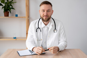 Portrait of a male doctor in the office working at the table. Serious doctor is waiting for the patient. Portrait of a smiling doctor in a bright office. Confident general practitioner ready to go.